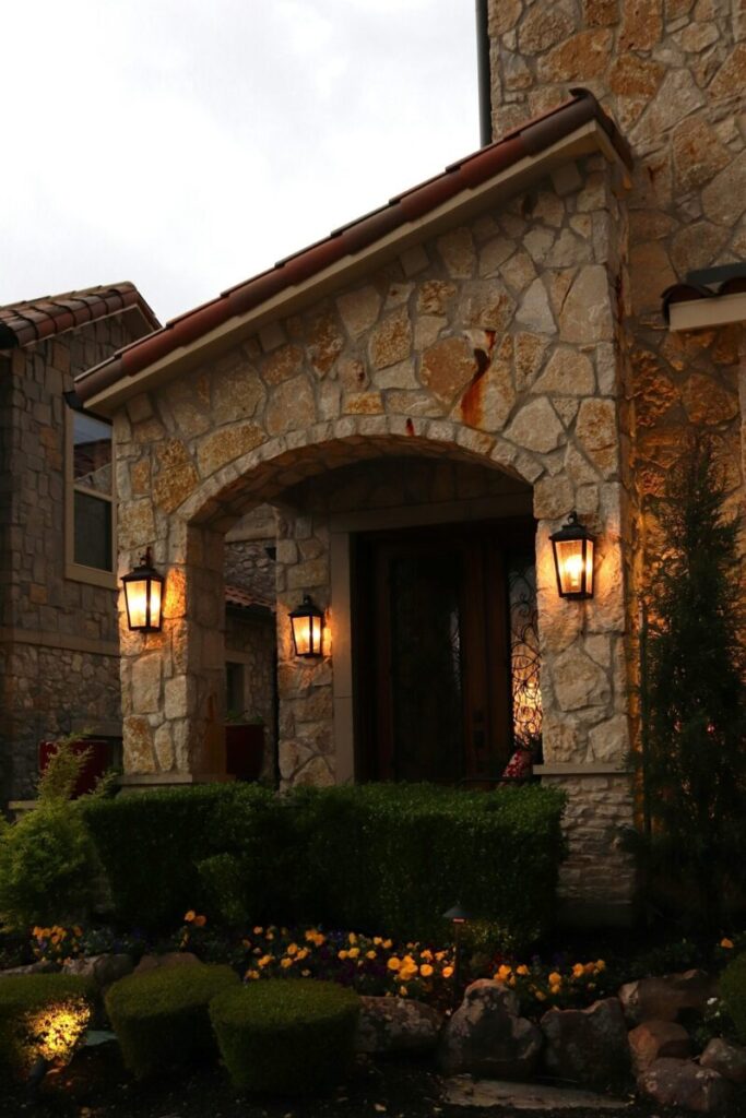 Charming stone entrance illuminated by classic lamps, surrounded by manicured greenery.